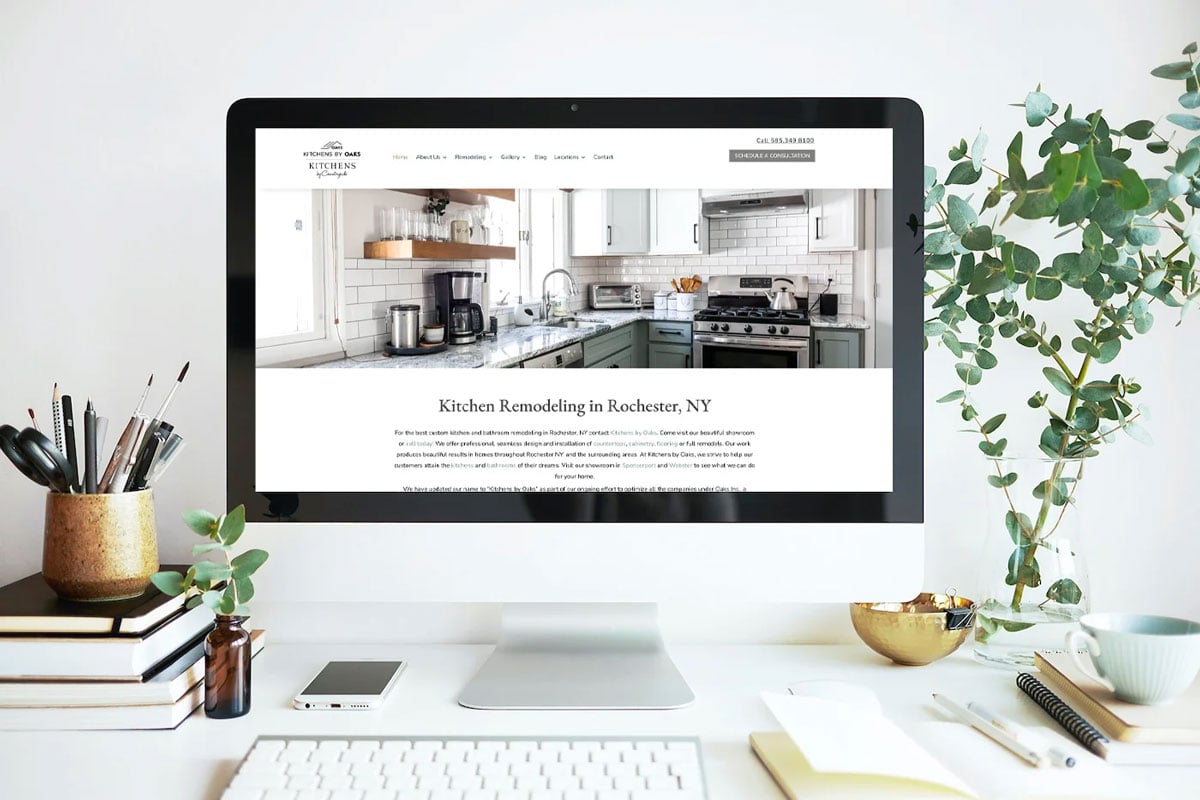 A modern home office workspace with a computer displaying a kitchen design webpage. The desk is organized with office supplies and a plant.