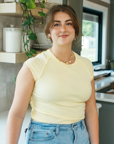 A young woman smiling in a kitchen with a plant and shelves. She is wearing a yellow shirt and jeans.