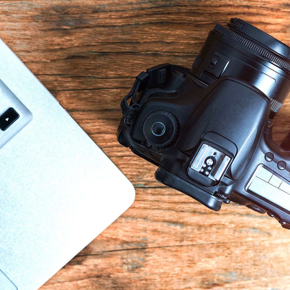A laptop and camera on a wooden desk. The camera has a leather strap.